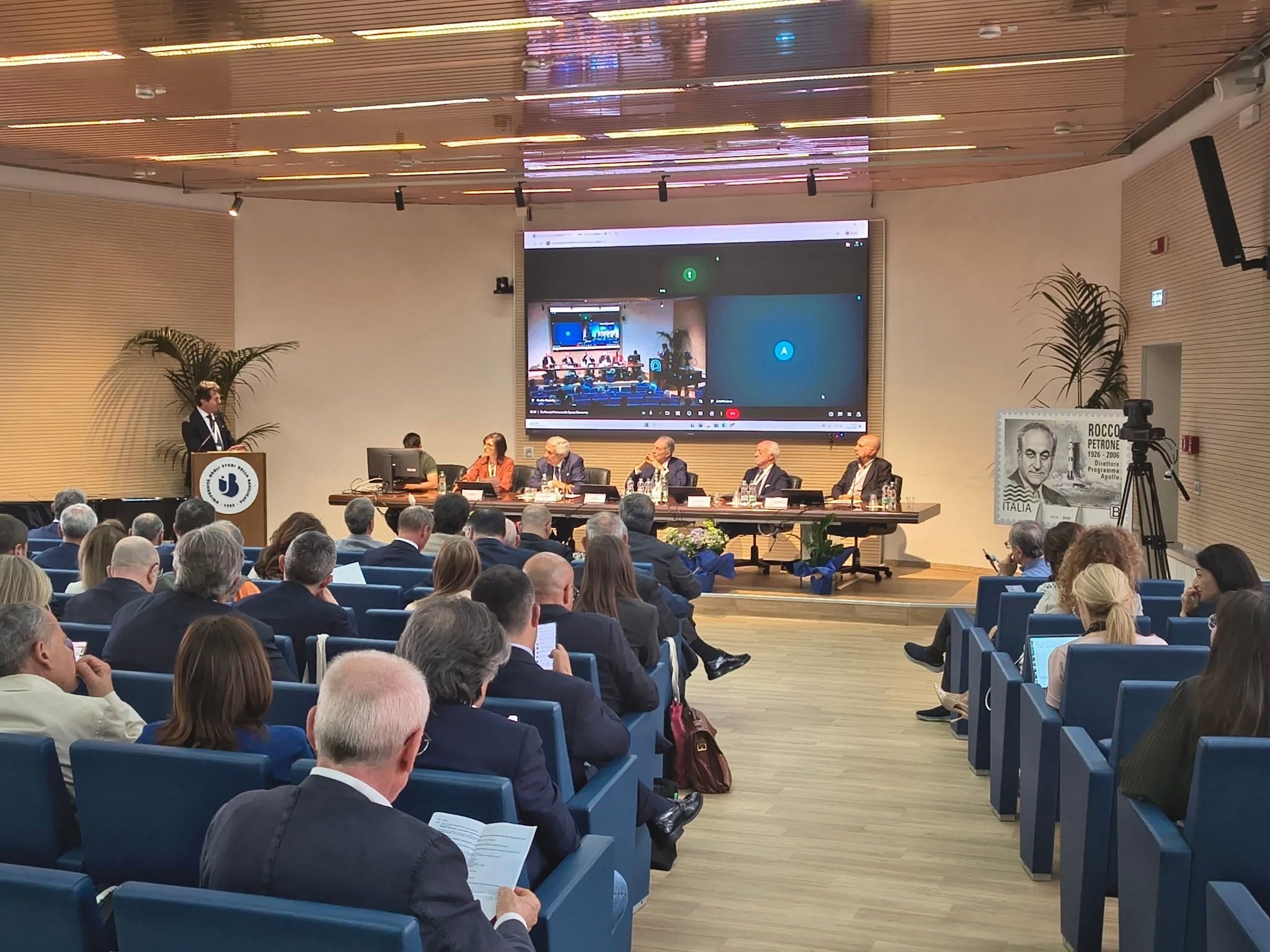 The hall of the University of Basilicata packed with participants during a conference session, with the speakers' table and the video link on screen